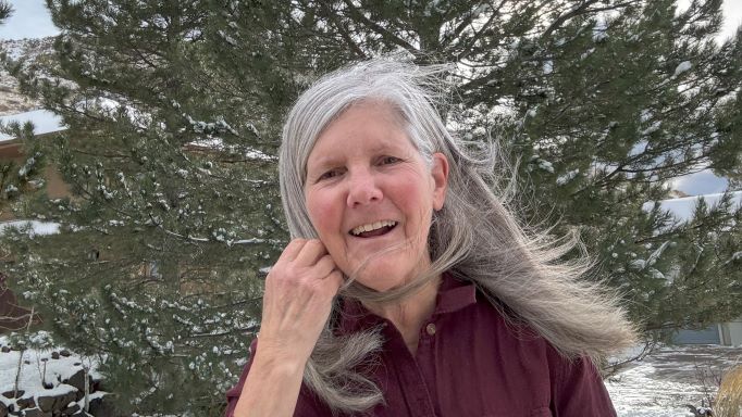 close-up of Nancy Goodman Torpey with her long silver hair blowing in the breeze standing in front of a large pine tree on a snowy day